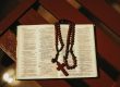 Otwarte Pismo Święte (Księga Psalmów, Psalmy 68–69) z różańcem z ciemnobrązowych drewnianych paciorków i drewnianym krzyżykiem, leżące na drewnianej ławce w ciepłym, przyciemnionym świetle.