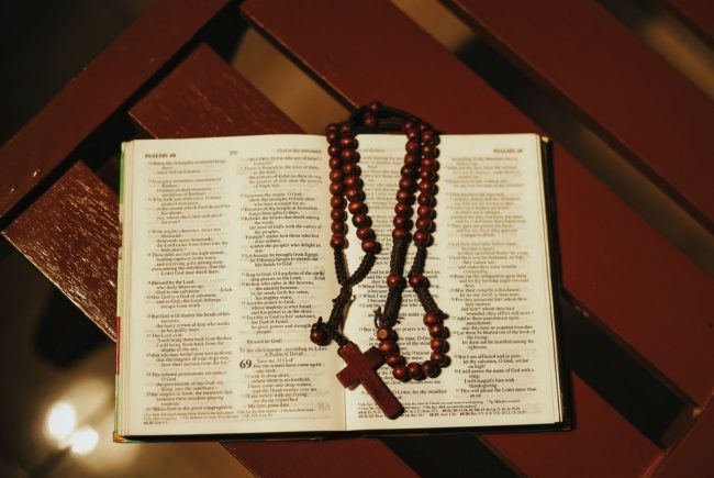 Otwarte Pismo Święte (Księga Psalmów, Psalmy 68–69) z różańcem z ciemnobrązowych drewnianych paciorków i drewnianym krzyżykiem, leżące na drewnianej ławce w ciepłym, przyciemnionym świetle.